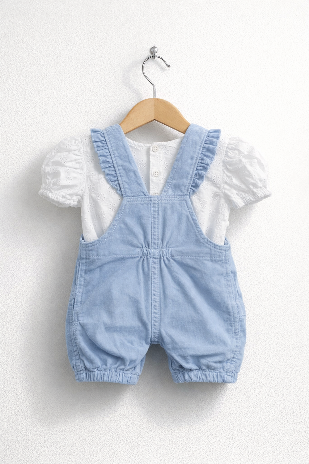 Light blue baby dungaree with ruffled straps paired with white embroidered top, neatly displayed on a wooden hanger against wall.